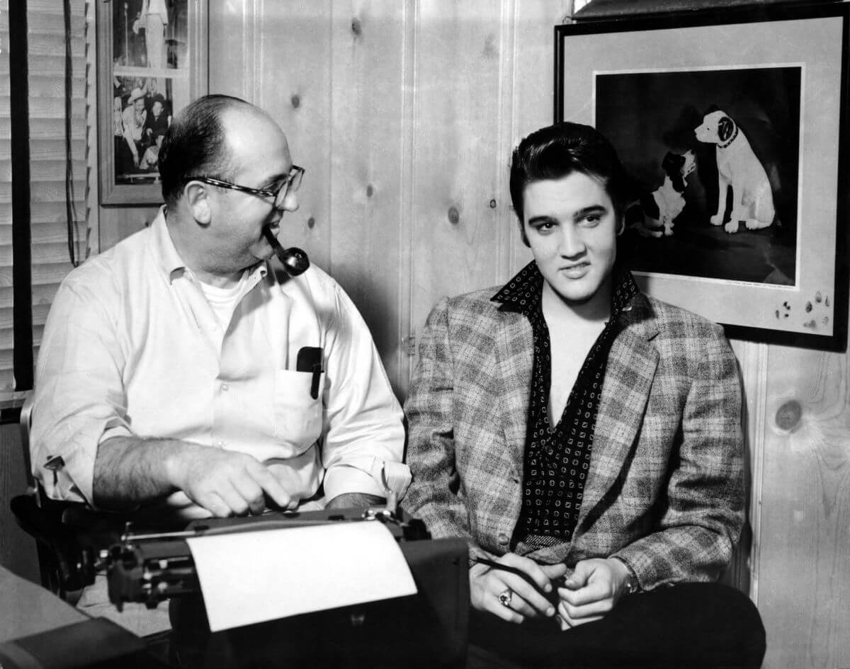 A black and white picture of Elvis sitting next to Colonel Tom Parker. Parker has a cigar in his mouth and sits at a typewriter.