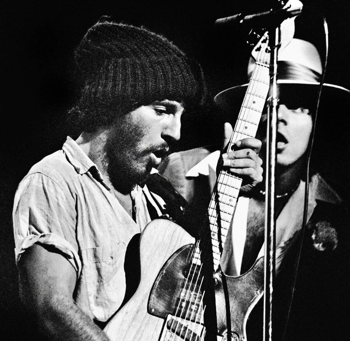 A black and white picture of Bruce Springsteen wearing a knit hat and playing guitar.