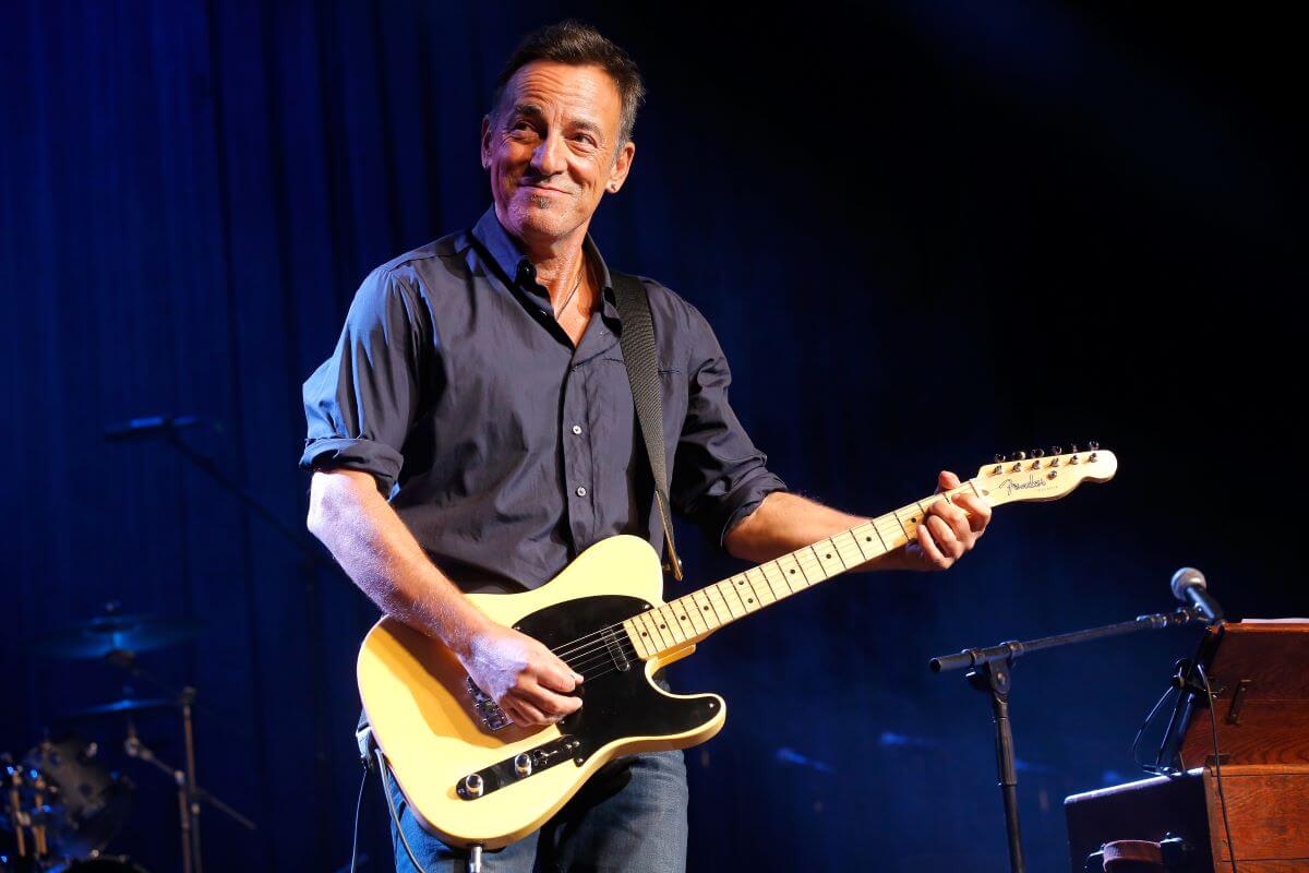 Bruce Springsteen wears a button down shirt and holds a guitar.