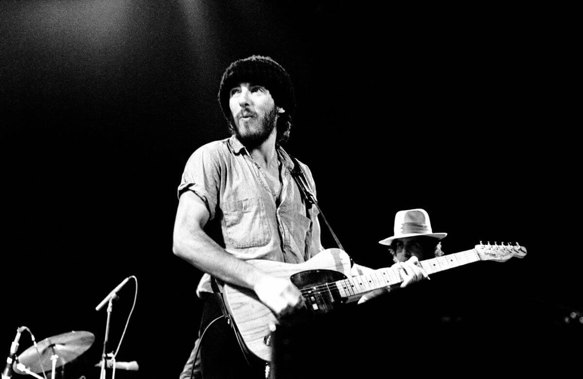 A black and white picture of Bruce Springsteen wearing a knit hat and strumming a guitar during a concert.