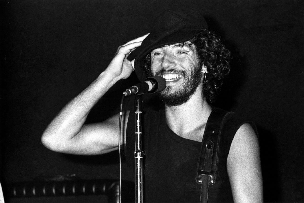 A black and white picture of Bruce Springsteen holding one hand up to his hat and standing in front of a microphone. He smiles.