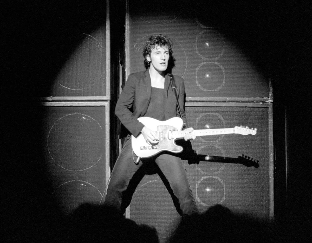 A black and white picture of Bruce Springsteen standing on stage in a spotlight. He holds a guitar.
