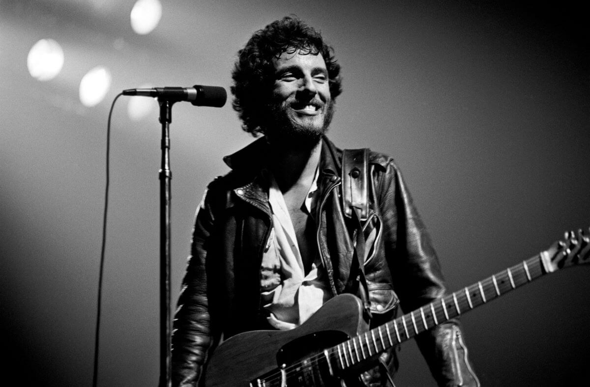 A black and white picture of Bruce Springsteen standing in front of a microphone with a guitar.