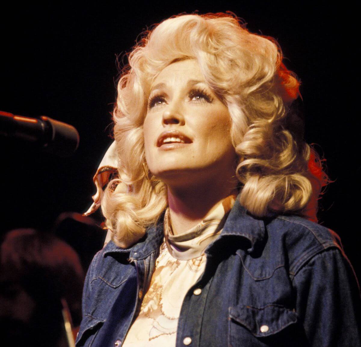 Dolly Parton wears a denim shirt and sits in front of a microphone. She looks up.
