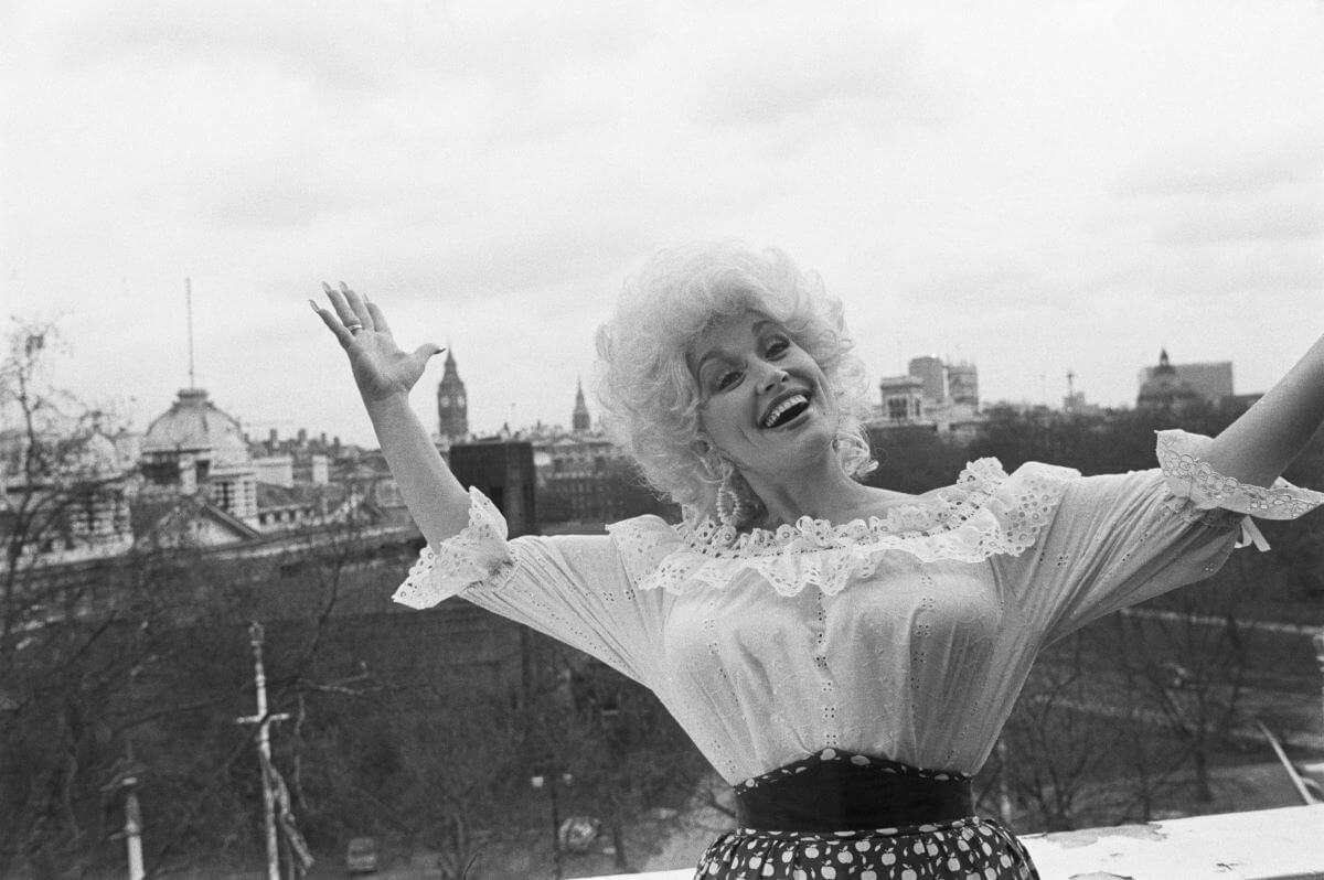 A black and white picture of Dolly Parton standing on a balcony and lifting her arms in the air.