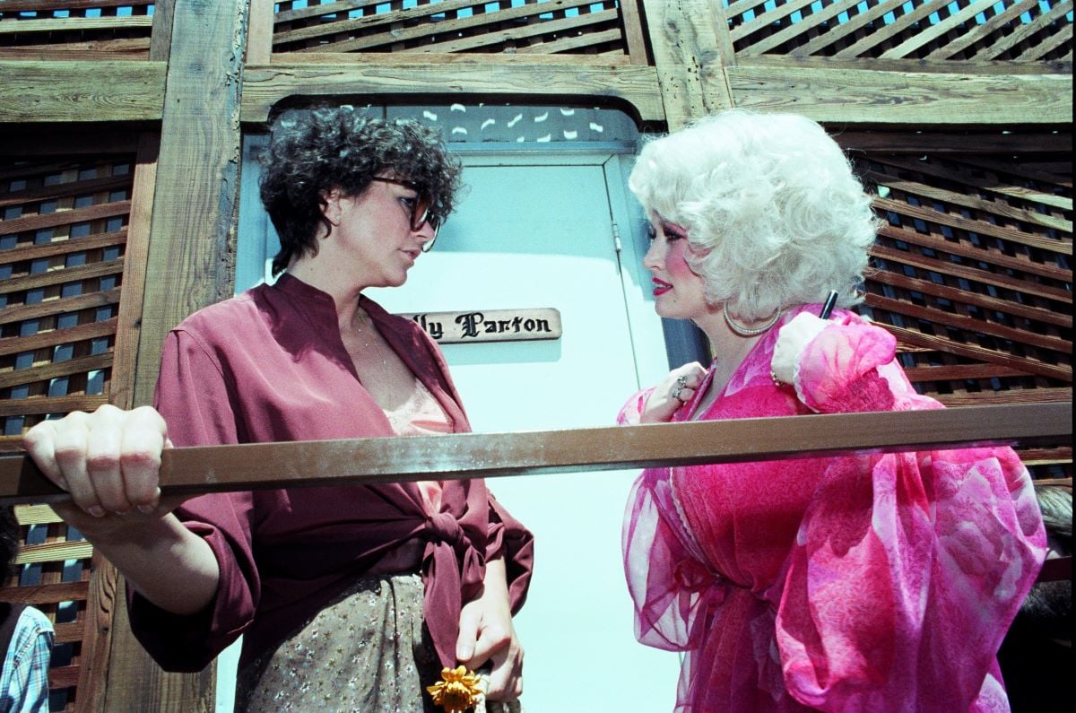 Linda Ronstadt and Dolly Parton stand on a wooden porch in front of a door with Parton's name on it.