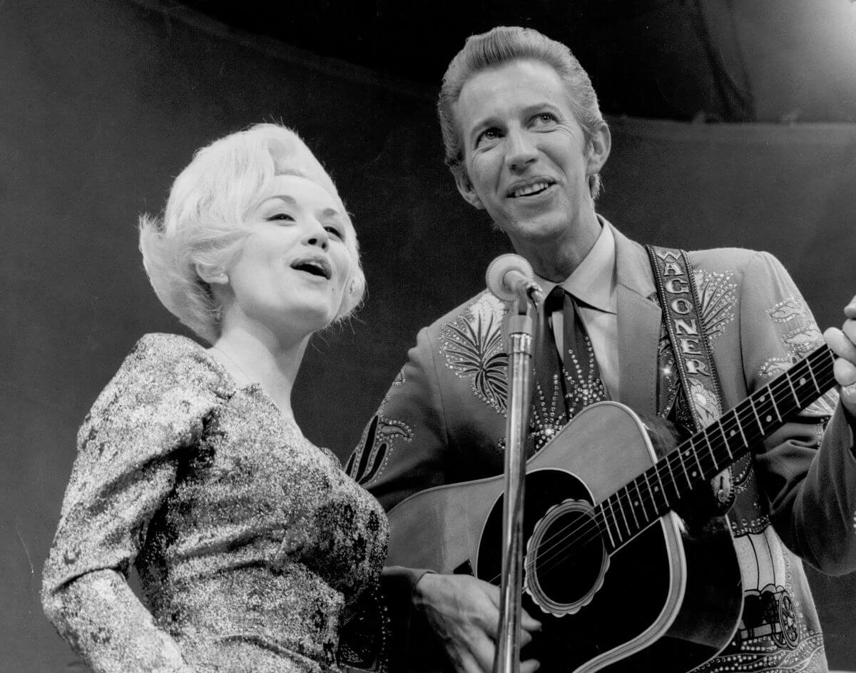 A black and white picture of Dolly Parton and Porter Wagoner singing into a microphone. He holds a guitar.