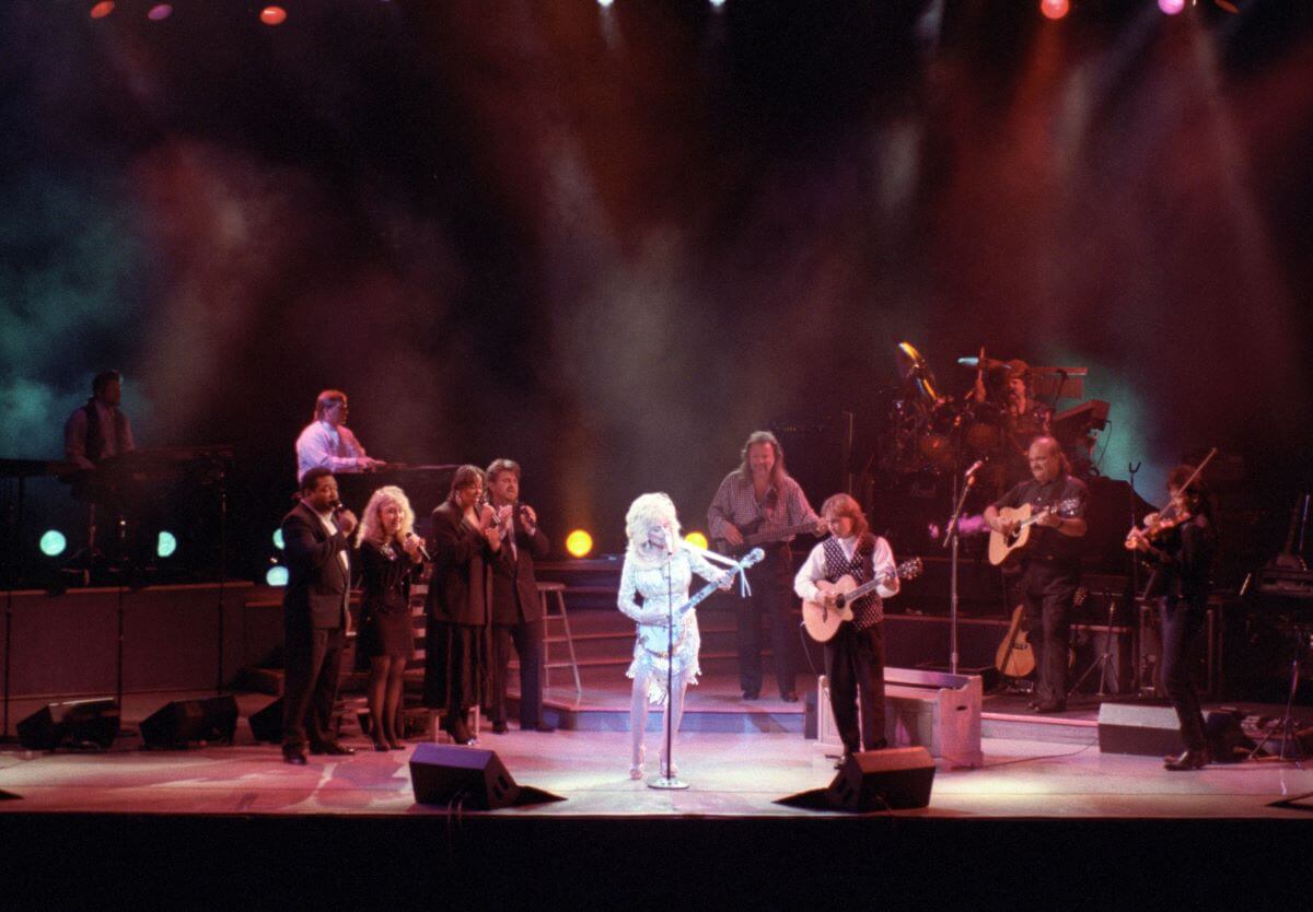 Dolly Parton wears white and stands in front of a microphone with her band.