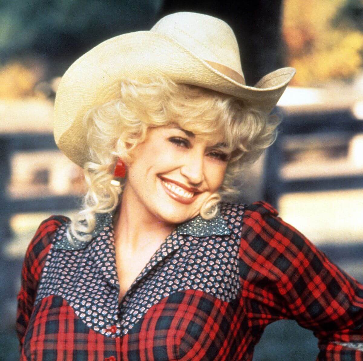 Dolly Parton wears a flannel shirt and a cowboy hat. She stands outside with one hand on her hip.