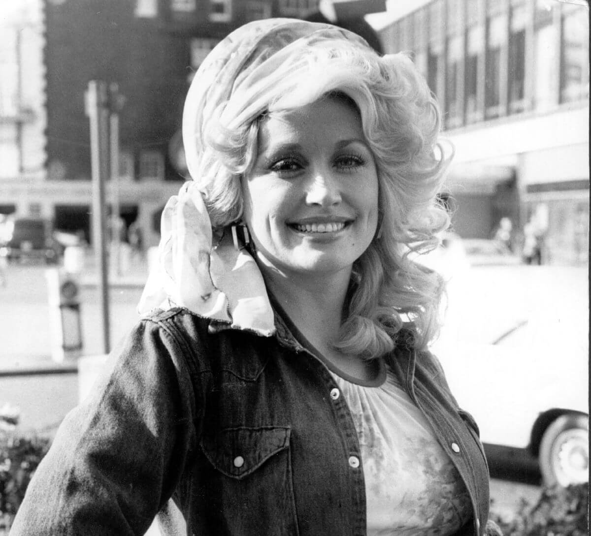 A black and white picture of Dolly Parton wearing a scarf around her hair and standing with one hand on her hip.