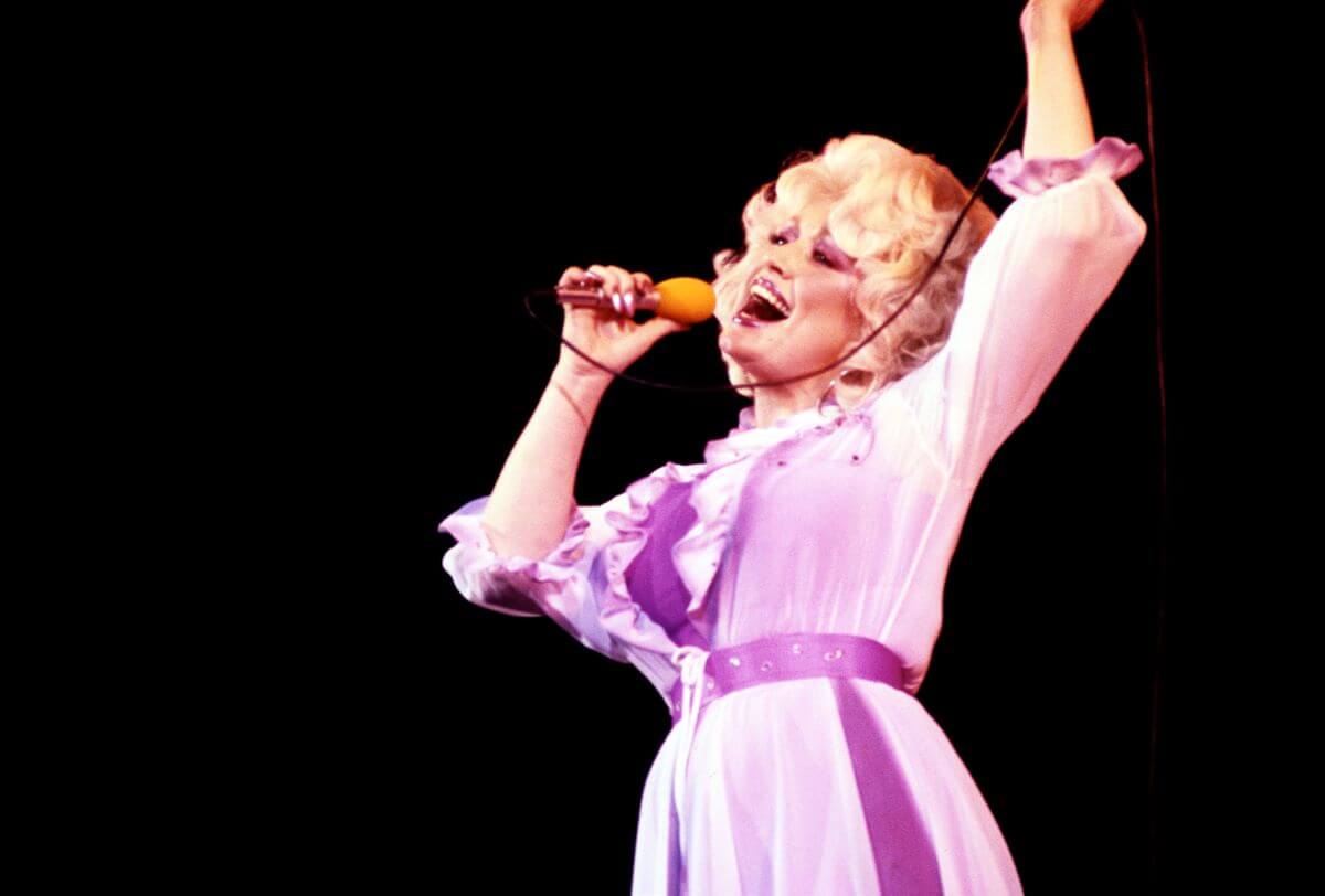 Dolly Parton wears a purple dress and sings into a microphone. She lifts one arm into the air.