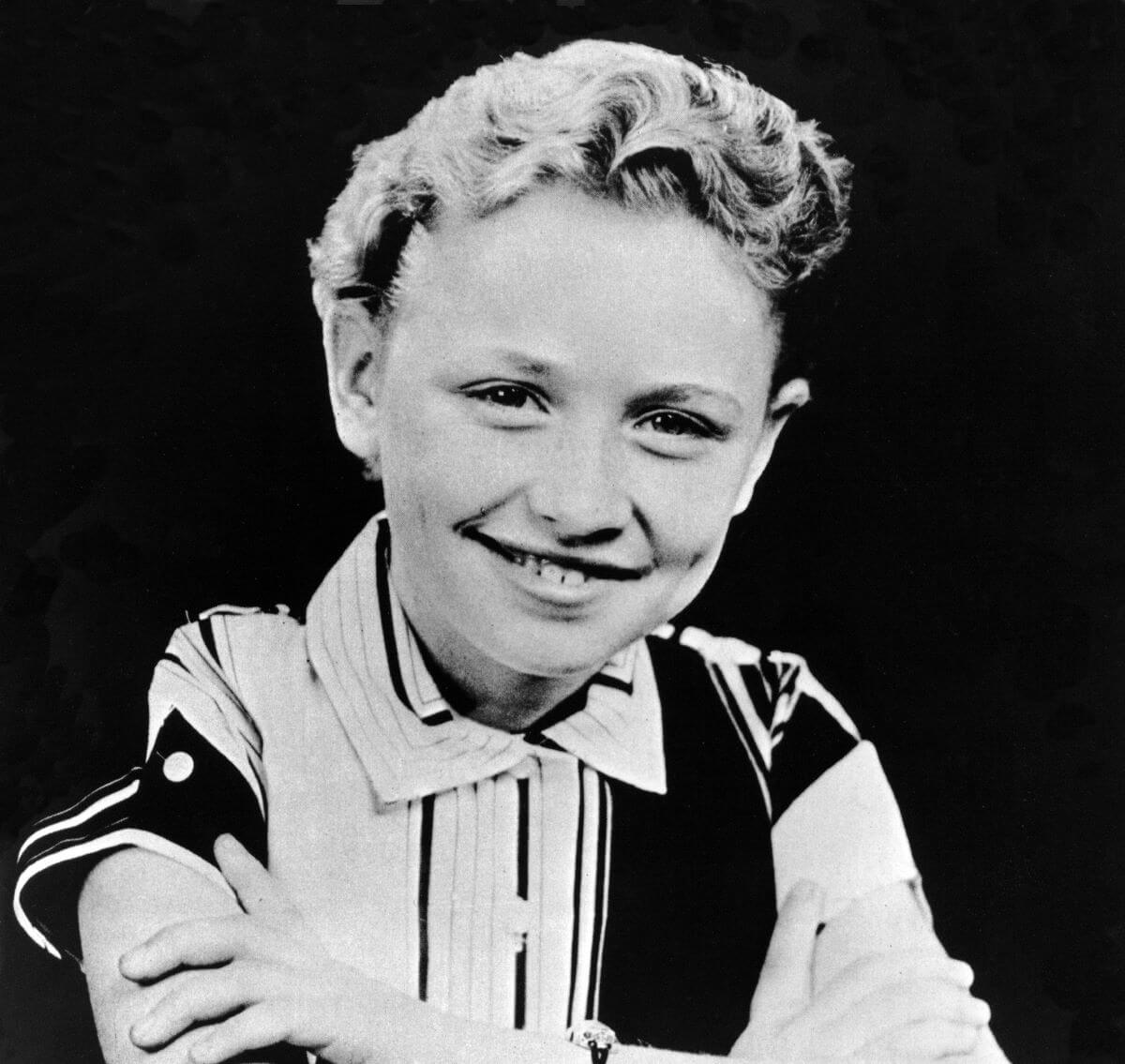 A black and white picture of Dolly Parton as a child. She wears a collared shirt and sits with her arms crossed.