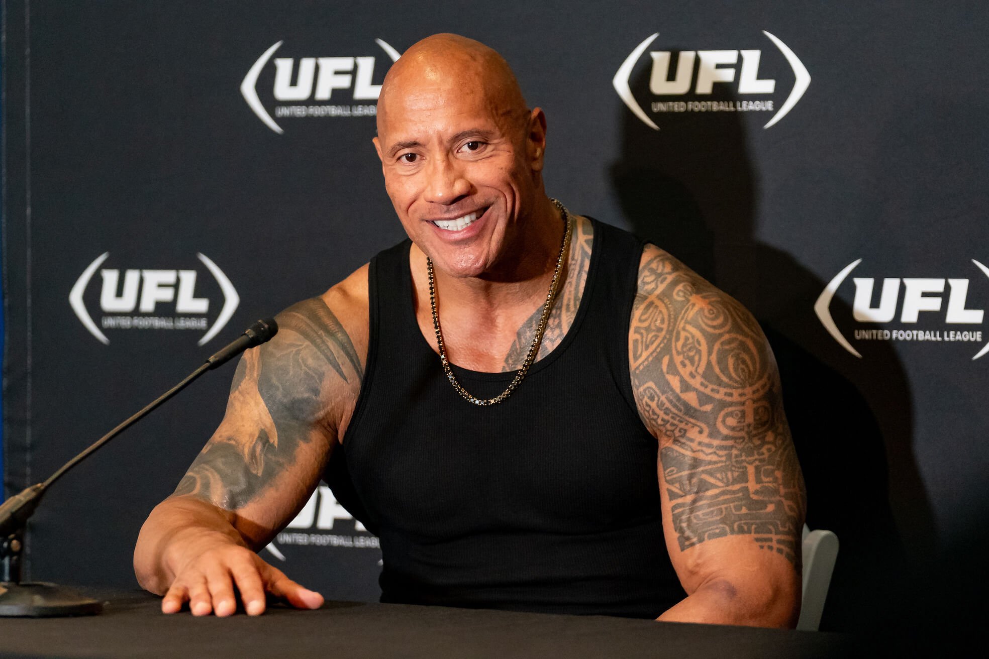 Dwayne 'The Rock' Johnson at a press conference. He's sitting at a table and wearing a black tank top that shows his tattoos.