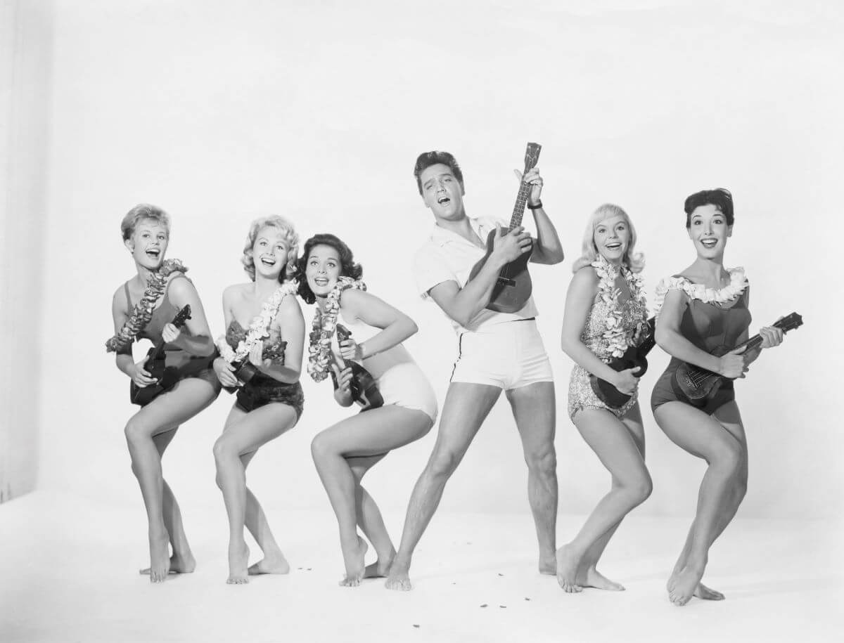 Elvis Presley holds a ukulele and poses with five women in leis who also hold ukuleles.