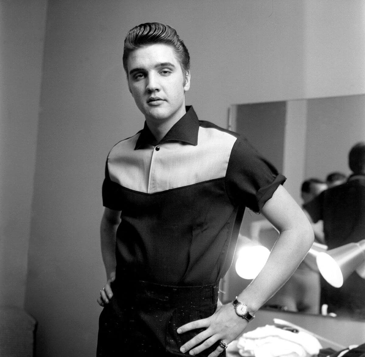 A black and white picture of Elvis Presley standing with his hands on his hips. He wears a collared shirt.