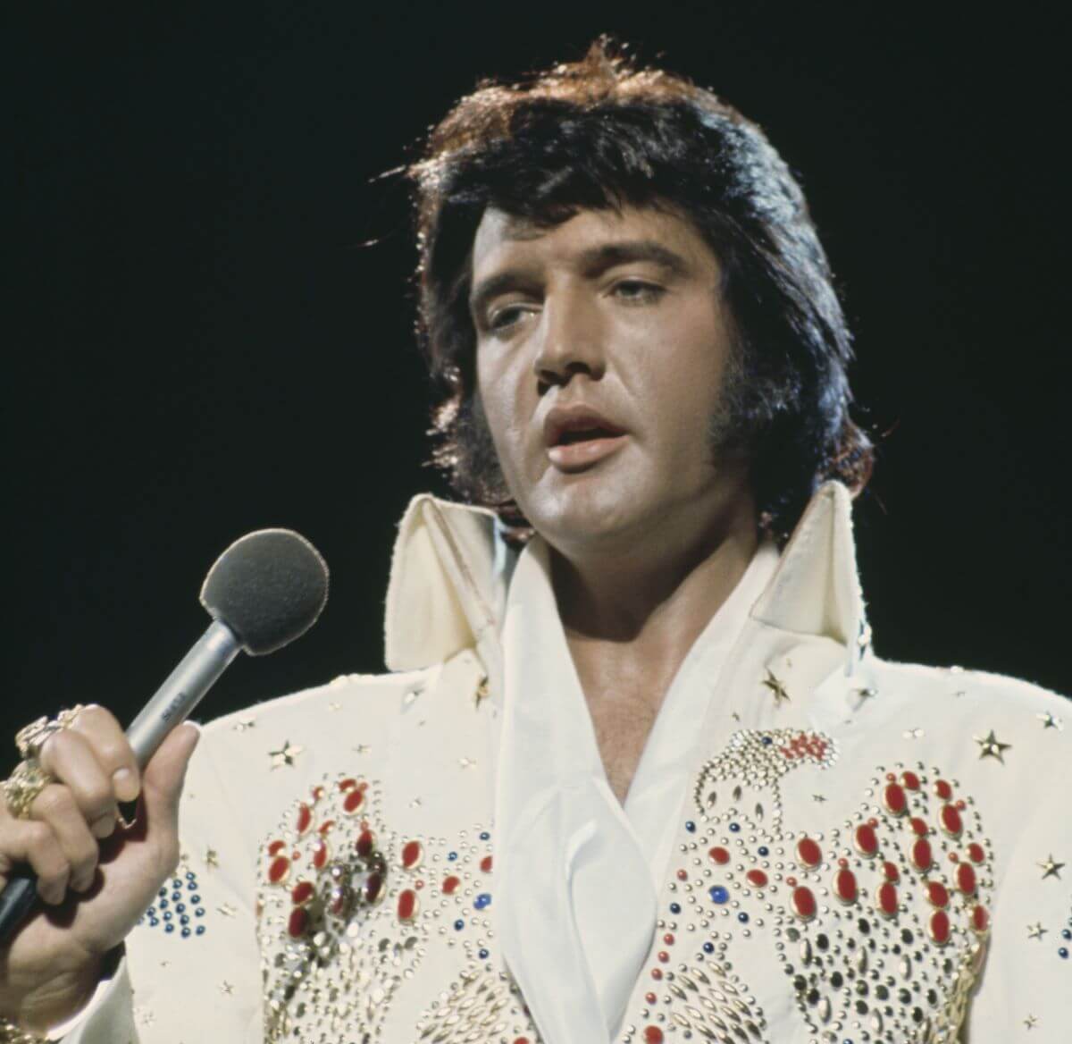 Elvis wears a white jumpsuit and holds a microphone.