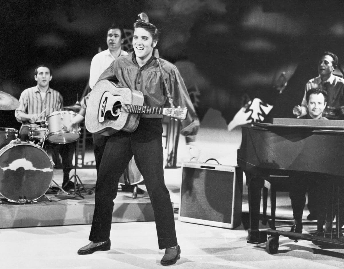 A black and white picture of Elvis standing in front of his band. He holds an acoustic guitar.