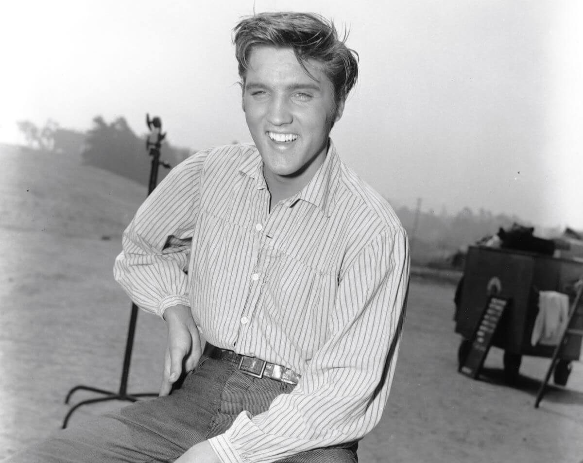 A black and white picture of Elvis sitting outside with one hand on his hip on the set of the film 'Love Me Tender.'
