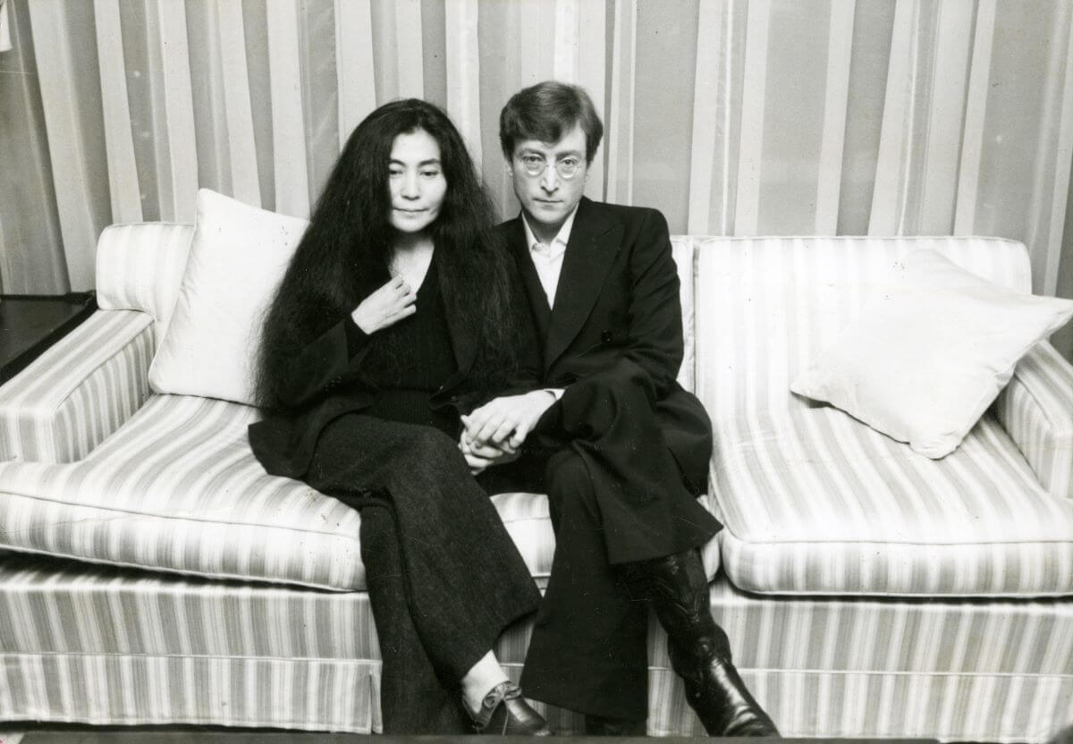 A black and white picture of Yoko Ono and John Lennon holding hands and sitting on a couch together.