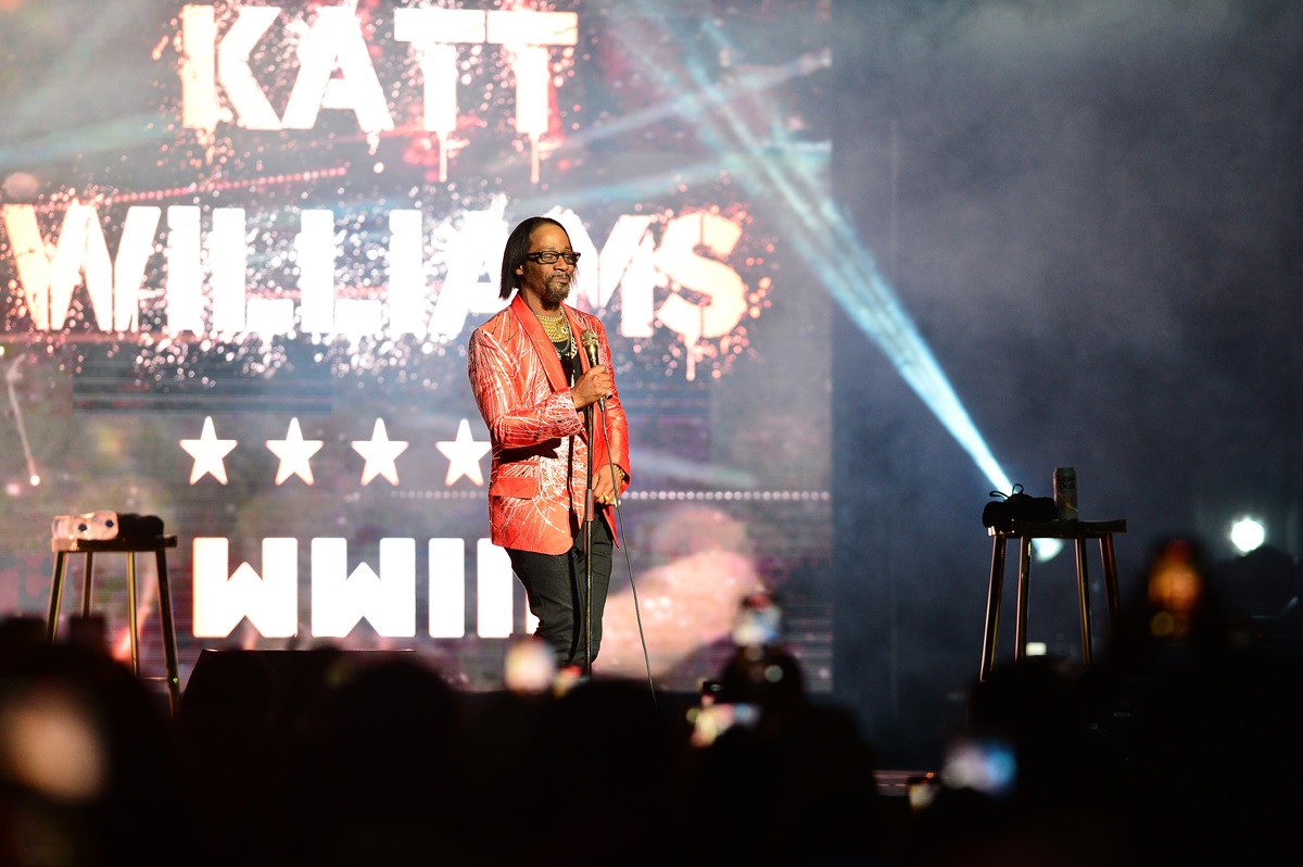 Katt Williams performing on stage in a red jacket and black jeans.