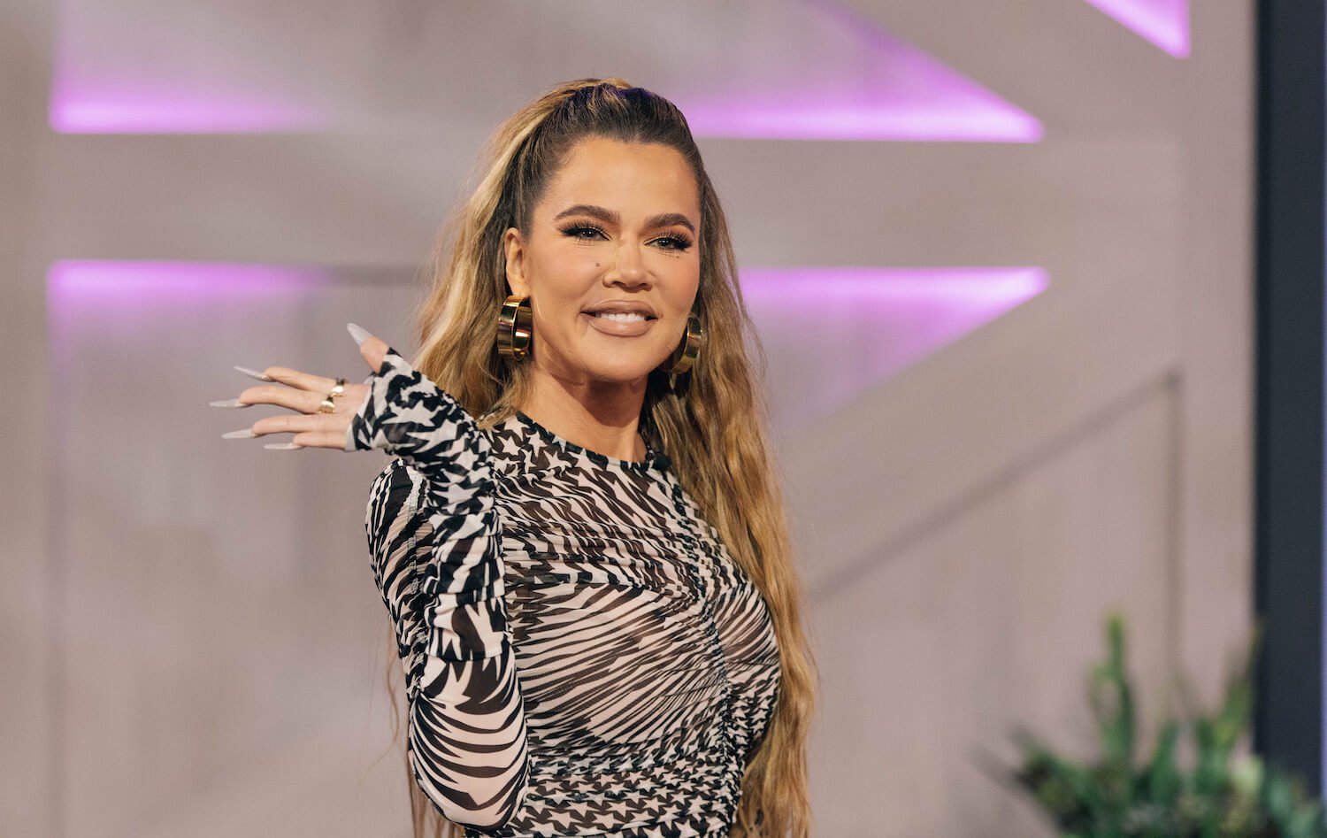 Khloé Kardashian smiling and waving before a talk show. Her hair is pulled back and she's wearing long sleeves.