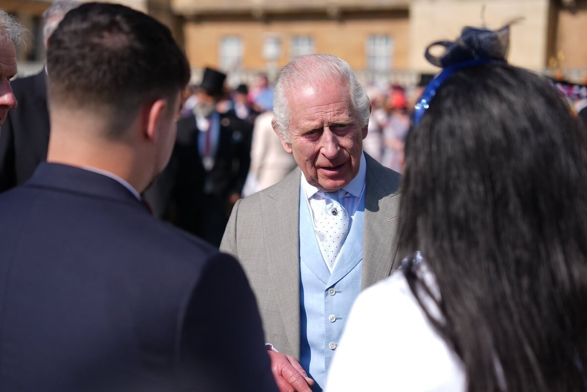 King Charles III, who can get 'points' by mentioning Prince Harry at Army ceremony, per a biographer, stands with two guests at a garden party