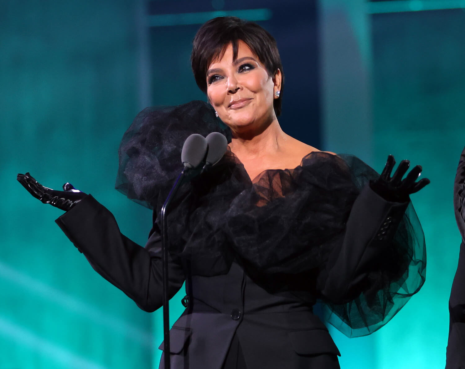 Kris Jenner shrugging and smiling. She's wearing a black dress with black gloves and is standing against a blue-green background.