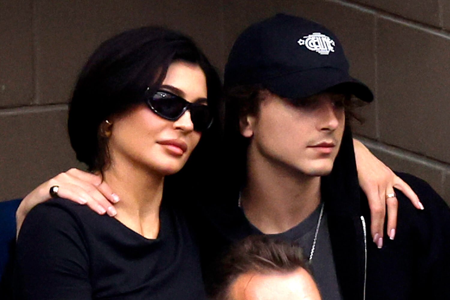 A close-up of Kylie Jenner wearing sunglasses and Timothée Chalamet wearing a hat at the US Open in 2023. They have their arms around each other.