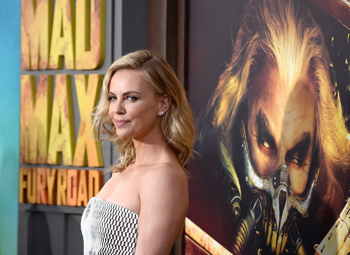 Charlize Theron posing in front of a 'Mad Max Fury Road' poster in a silver dress.