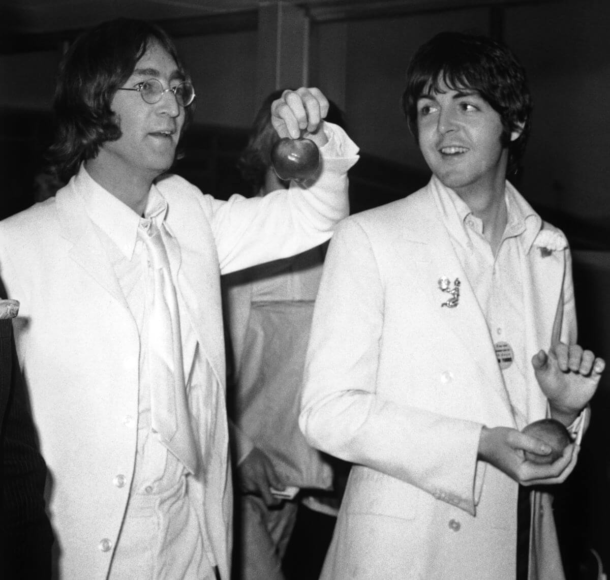 A black and white picture of Paul McCartney and John Lennon walking together. They both wear white suits and hold apples.