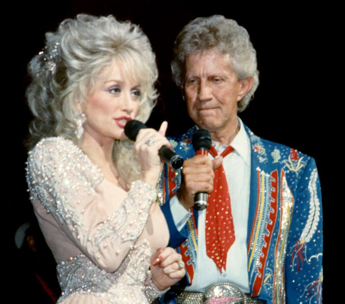 Porter Wagoner holds a microphone and watches Dolly Parton, who speaks into a microphone.