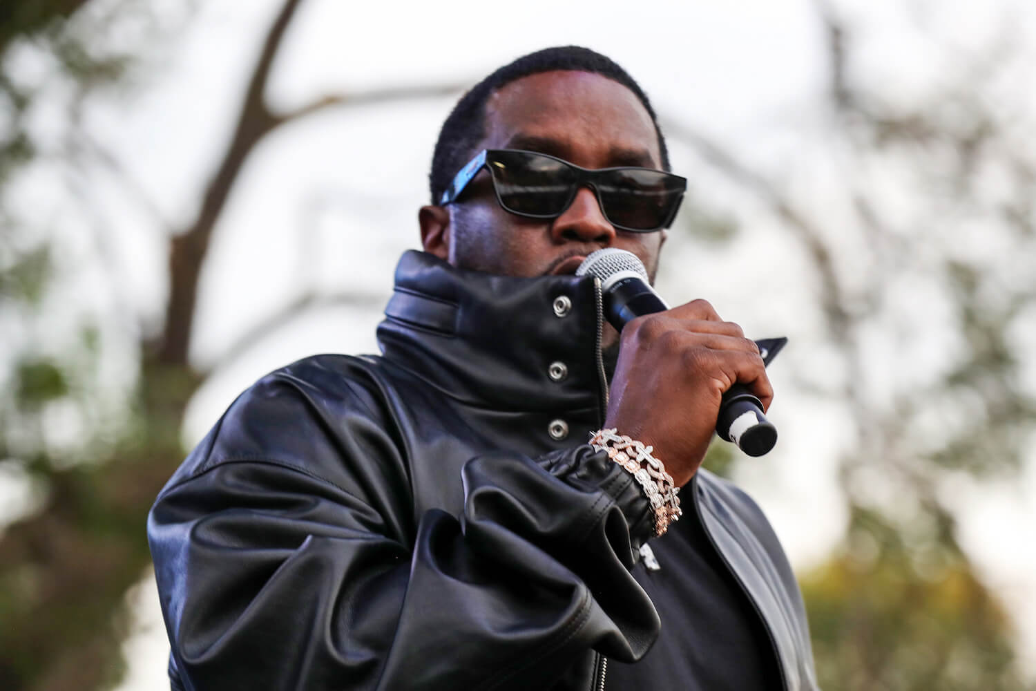 Sean 'P. Diddy' Combs holding a microphone up to his mouth while outside. He's wearing a leather jacket and sunglasses.