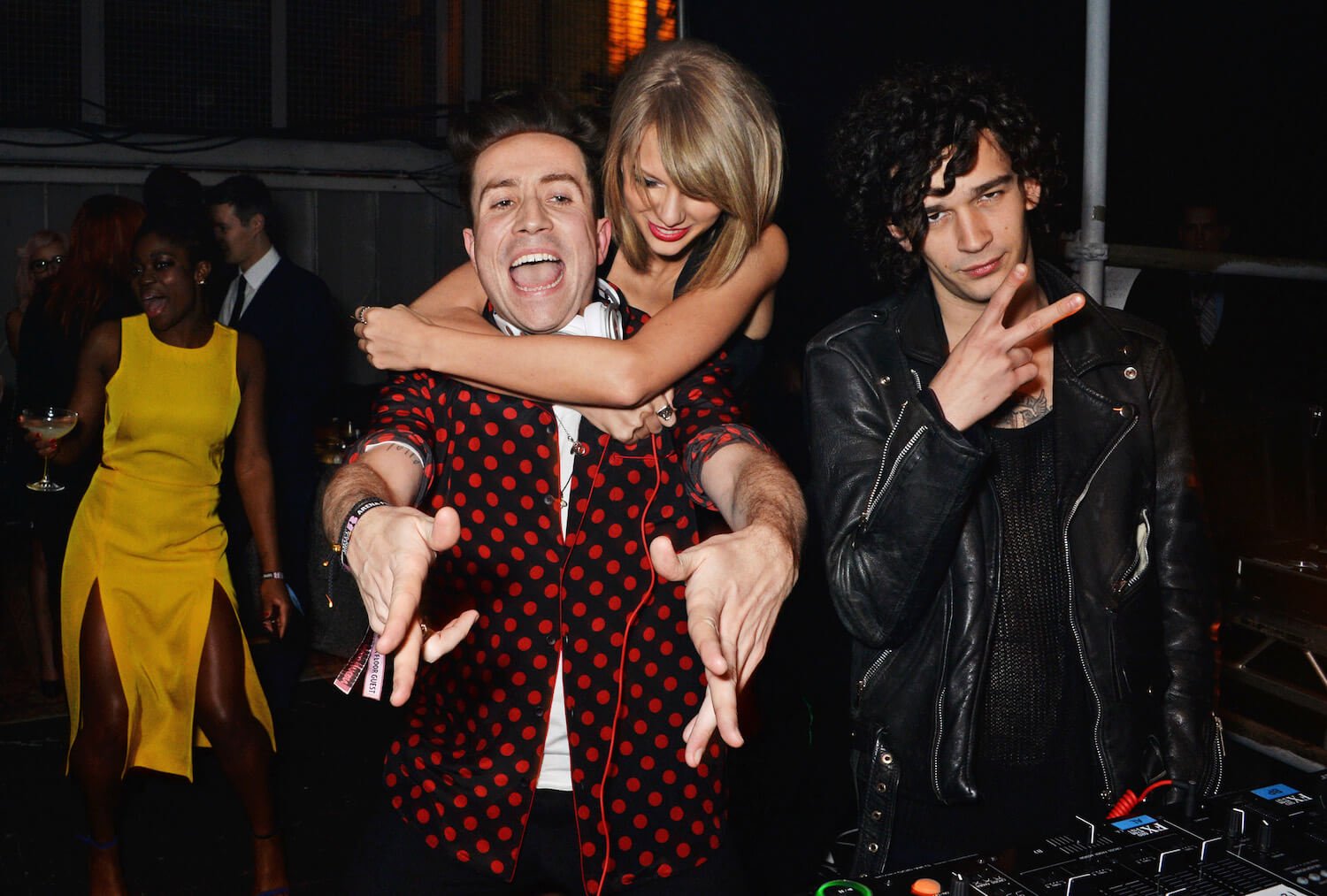 Taylor Swift with her arms around Nick Grimshaw as he's reaching forward and smiling. Matty Healy is next to them.