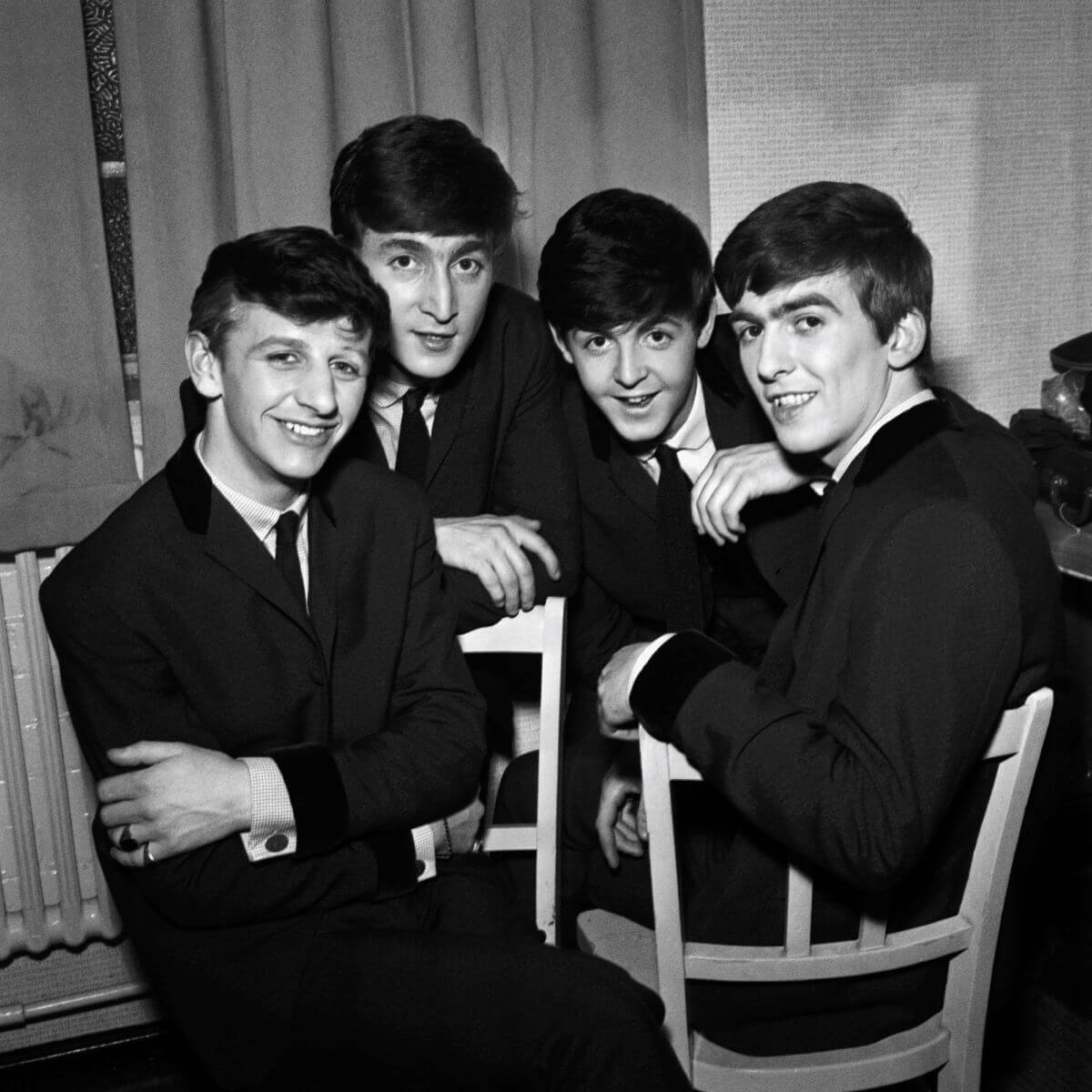 A black and white picture of Ringo Starr, John Lennon, Paul McCartney, and George Harrison of The Beatles gathered together on chairs. They wear suits.