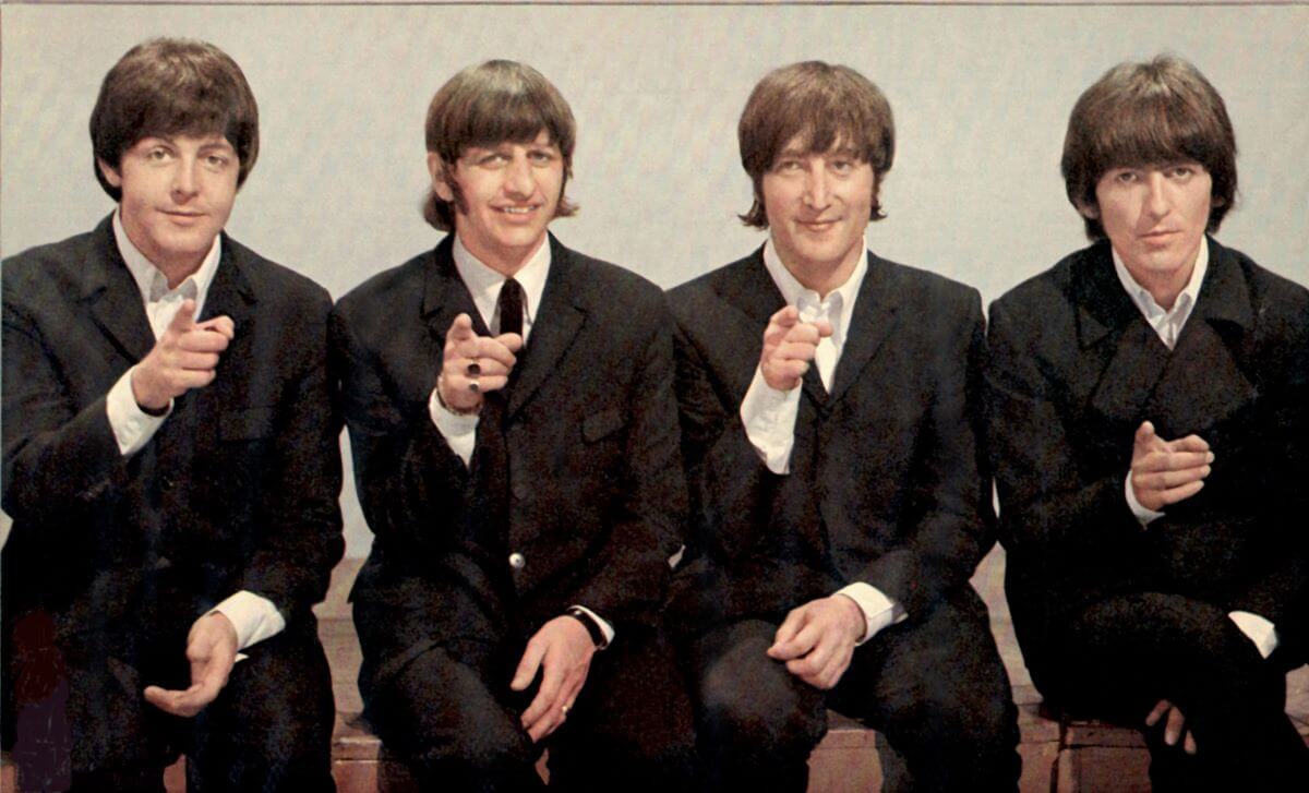 The Beatles wear suits and sit in a row. They each point one hand at the camera.