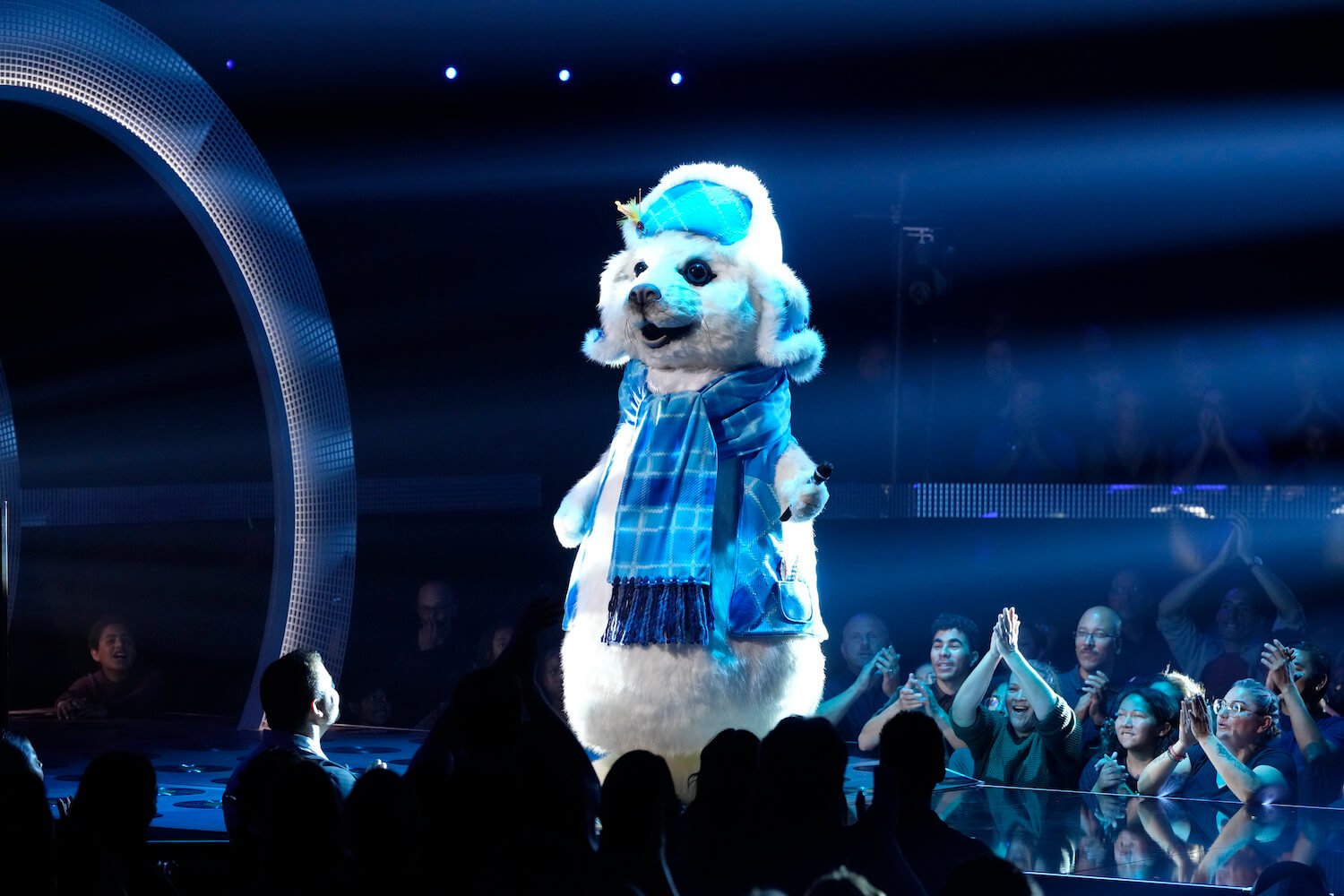 A man singing in a white seal costume against a dark background while surrounded by a sitting audience in 'The Masked Singer' Season 11