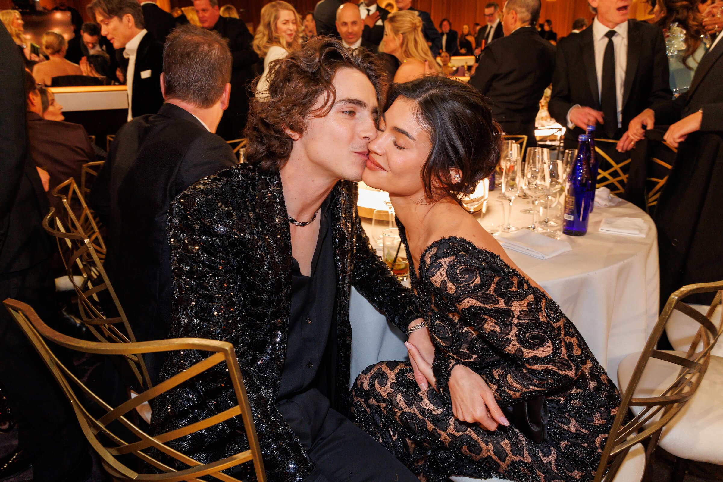 Timothée Chalamet kissing Kylie Jenner on the cheek as they sit at a table at the 81st Golden Globe Awards. He is wearing a low-cut black suit and she is wearing a long-sleeve black and tan dress.