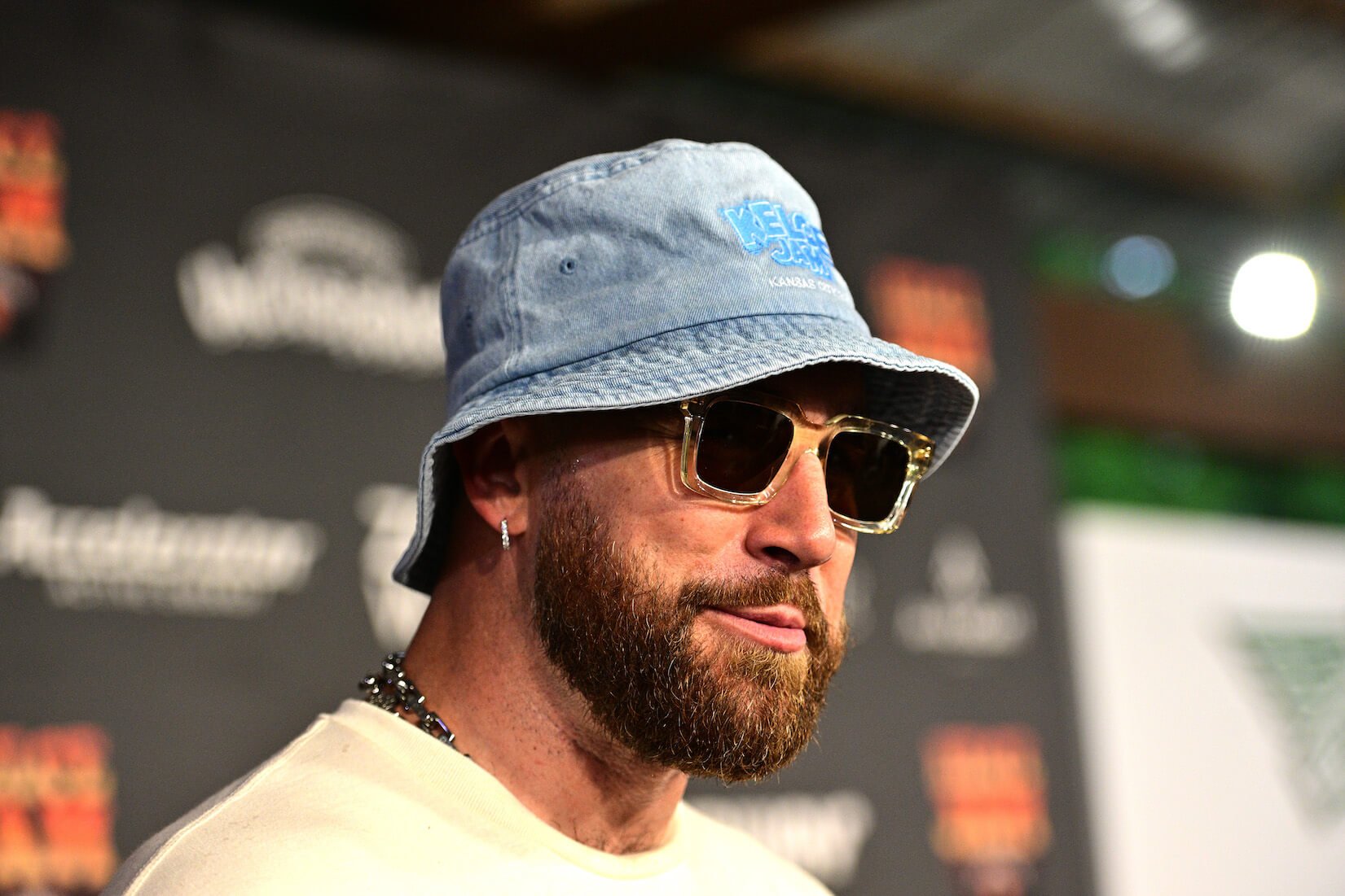 A close-up of Travis Kelce wearing sunglasses and a denim bucket hat at an event