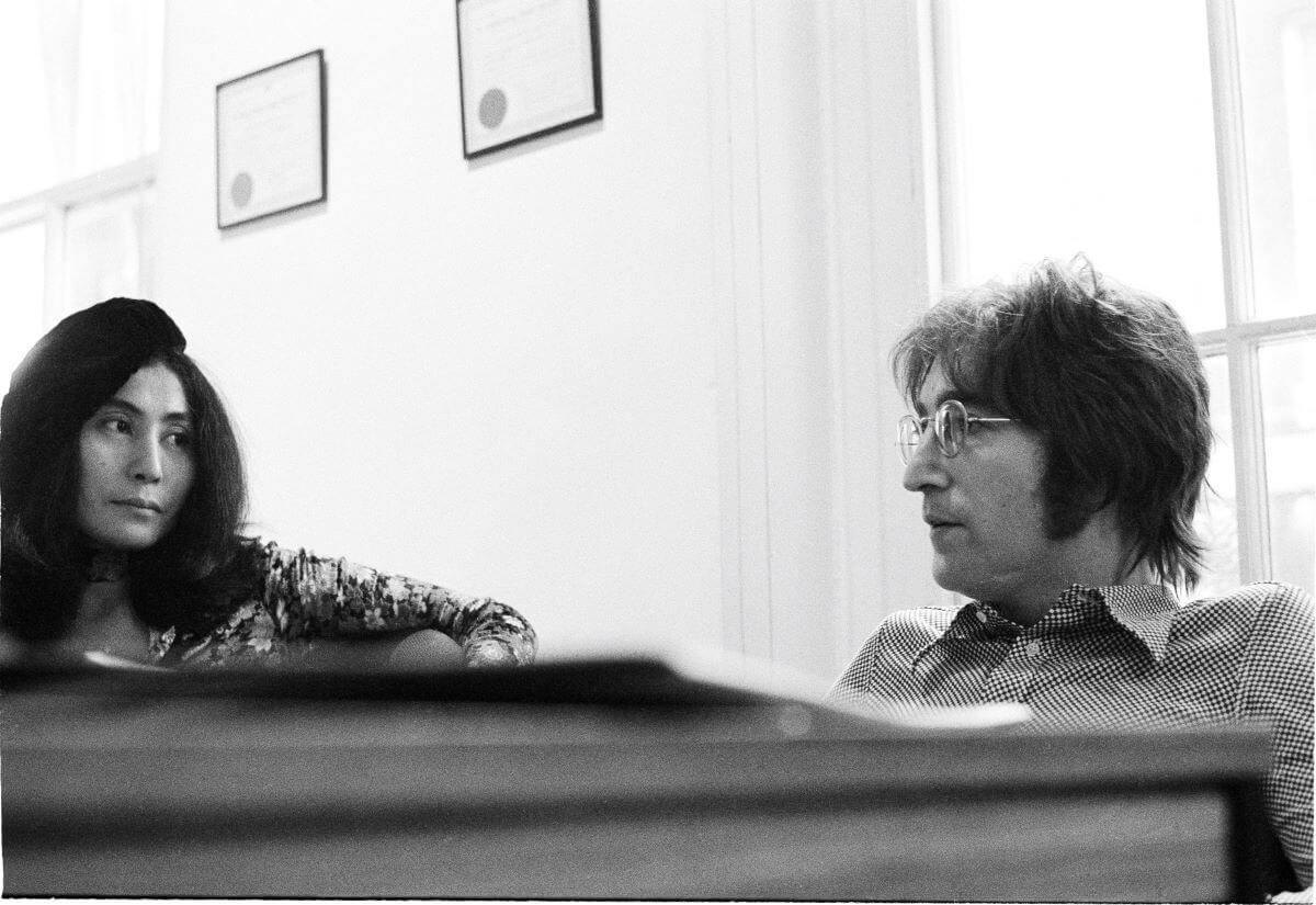 A black and white picture of Yoko Ono and John Lennon sitting on a couch together. She looks at him.