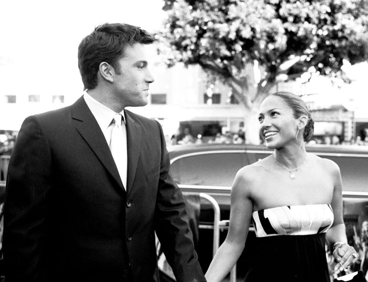 A black and white picture of Ben Affleck and Jennifer Lopez holding hands. They stand in front of a car.