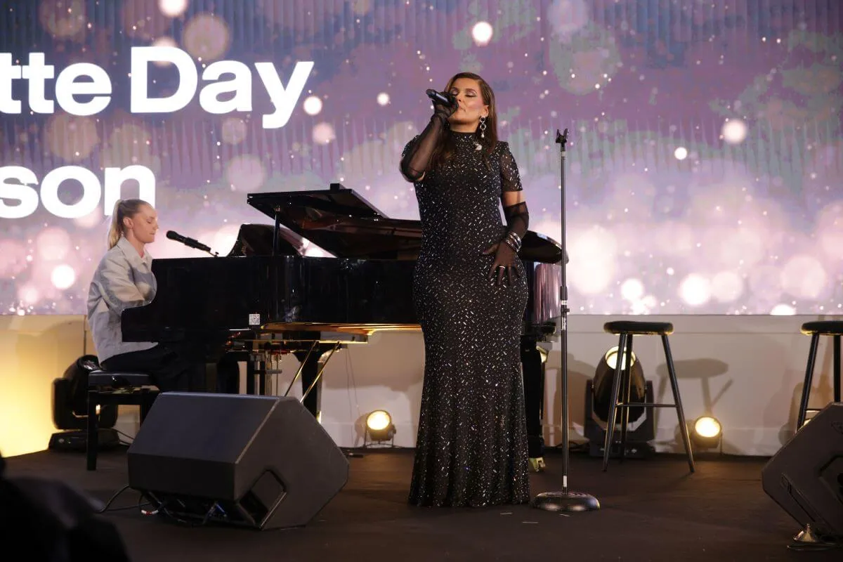 Nelly Furtado wears a long black dress and sings into a microphone. She stands in front of a piano.