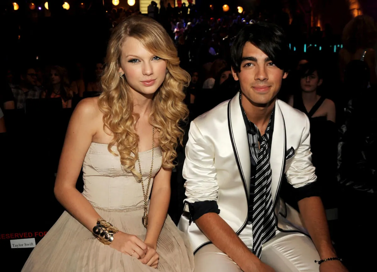 Taylor Swift wears a tan dress and sits next to Joe Jonas, who wears a white jacket.