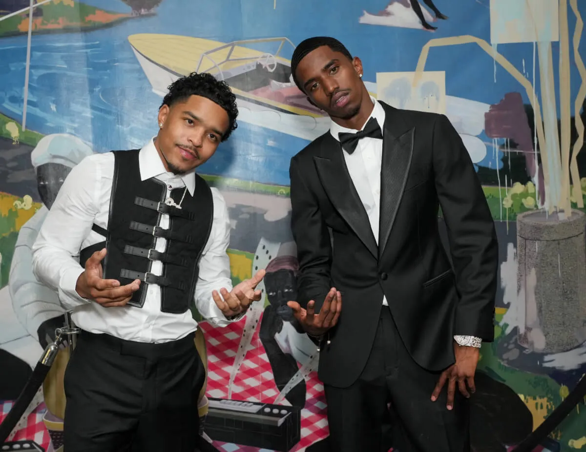 Sean 'Diddy' Combs' sons, Justin and Christian Combs, stand in front of a painting. Justin wears a dress shirt and a vest. Christian wears a tuxedo.