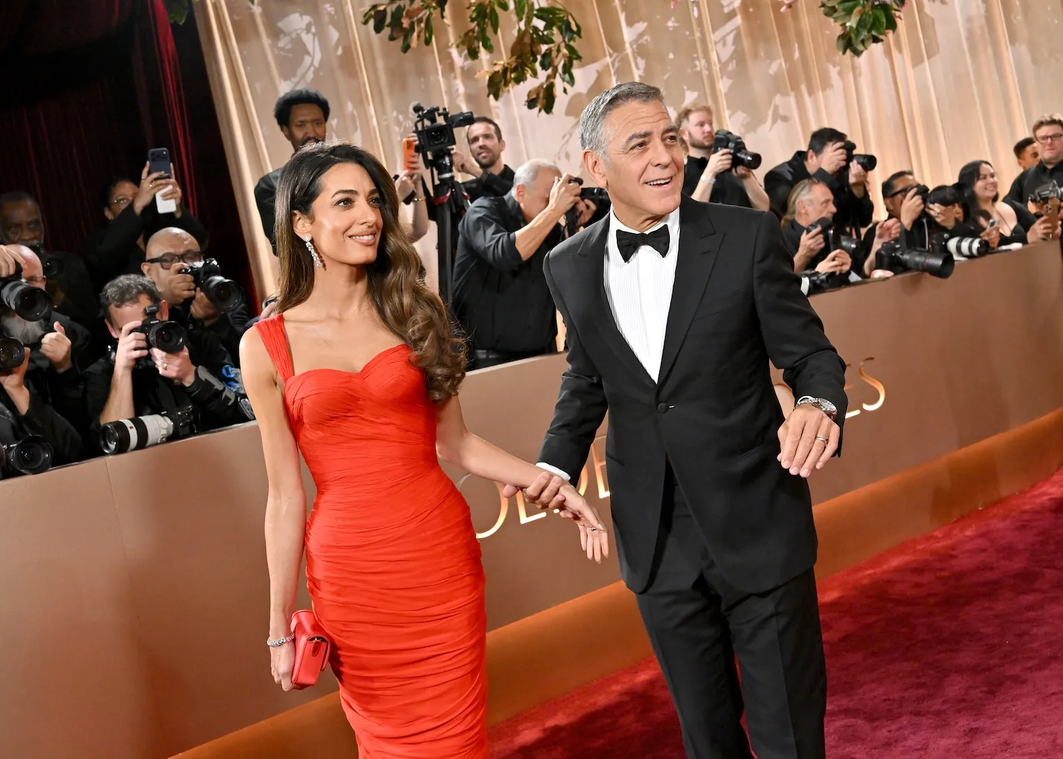 Amal Clooney in a red dress and George Clooney in a tuxedo at the 2026 Golden Globes. They're holding hands on the red carpet.