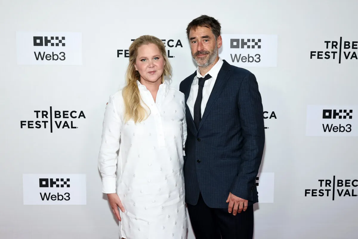 Amy Schumer wears a white dress and stands with her husband, Chris Fischer. He wears a suit.