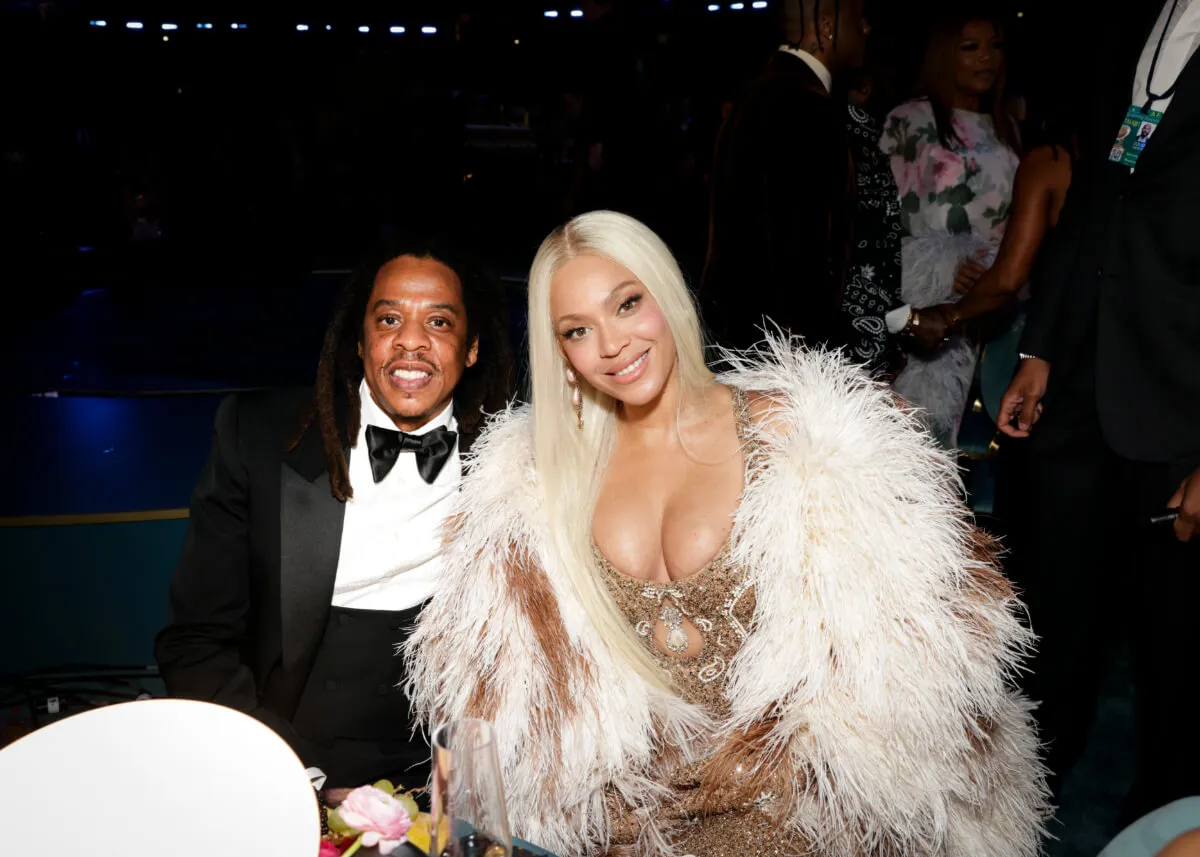 Jay-Z wears a tuxedo and sits with Beyonce, who wears a gold dress with a white fur jacket.