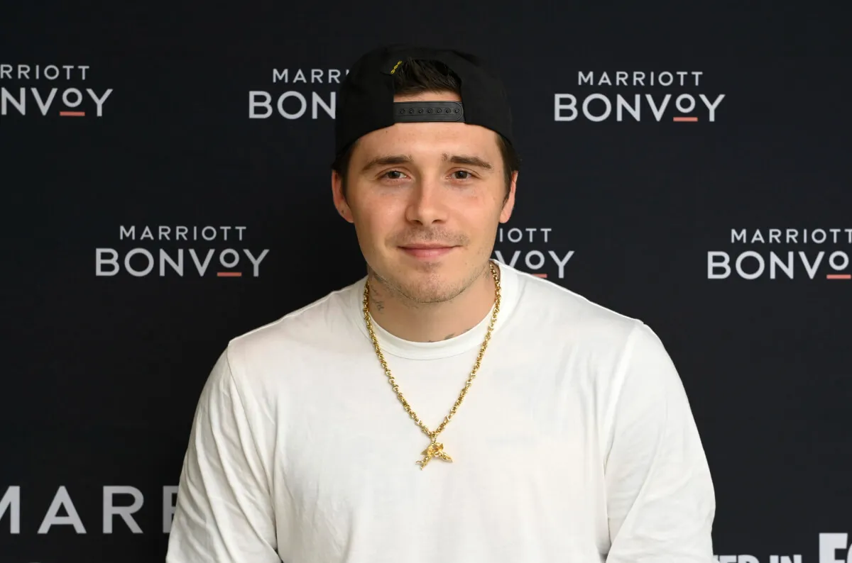 Brooklyn Beckham wears a white shirt with a gold necklace and a backwards baseball hat.