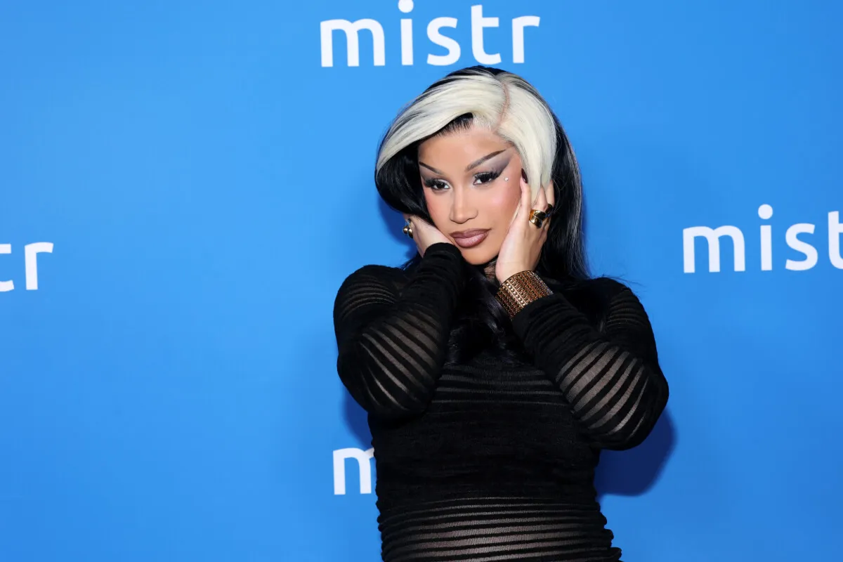 Cardi B wears black and touches her face. She stands in front of a blue background.