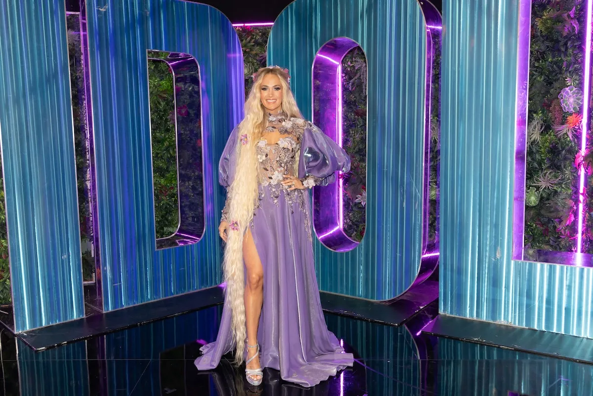 Carrie Underwood on 'American Idol' in 2025. She's posing with her hand on her hip and wearing a long purple dress with a thigh-high slit.