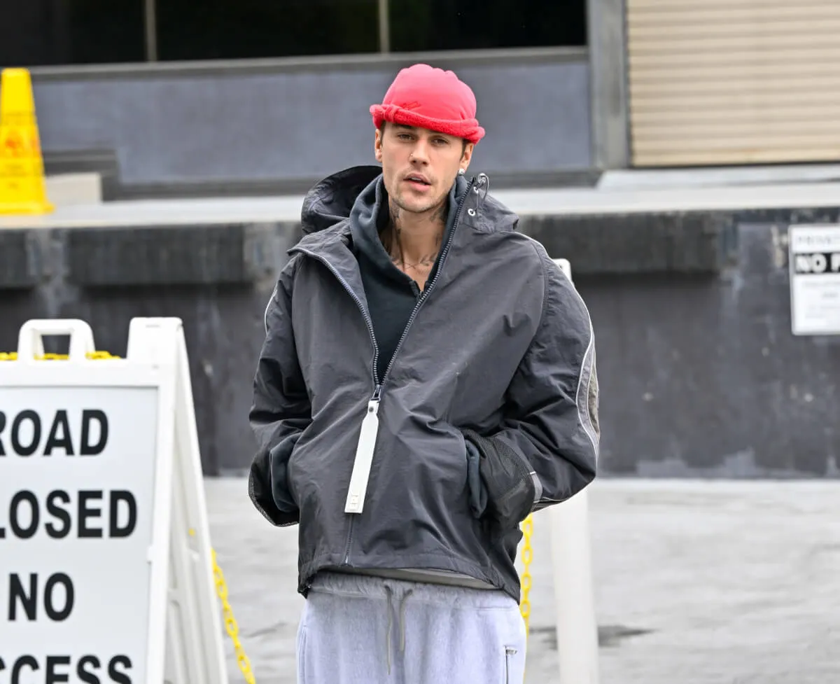 Justin Bieber wears a pink hat and black leather jacket. He stands in front of a parking lot.